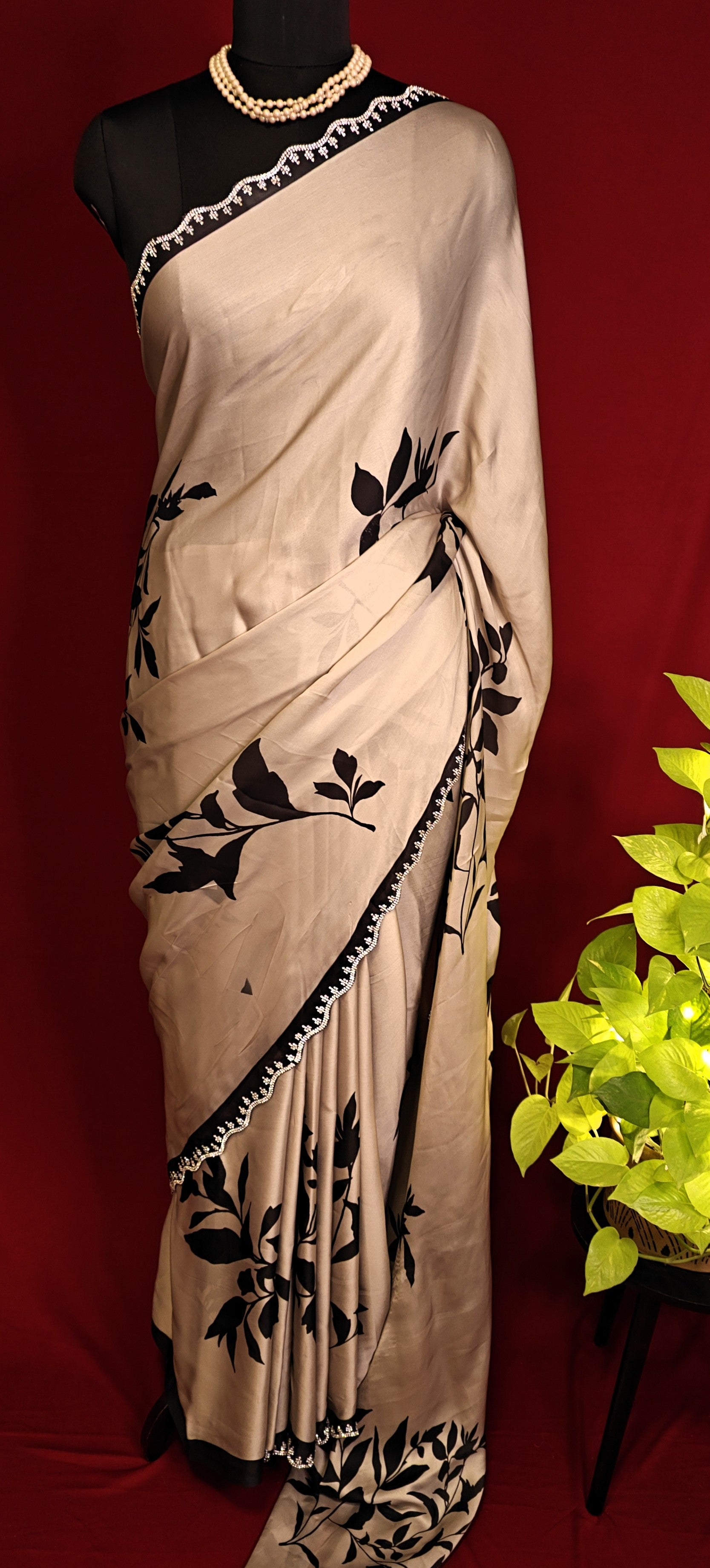 Cream satin georgette saree with medium-sized black leaf print, black motif border, and matching blouse piece on a mannequin against a red background