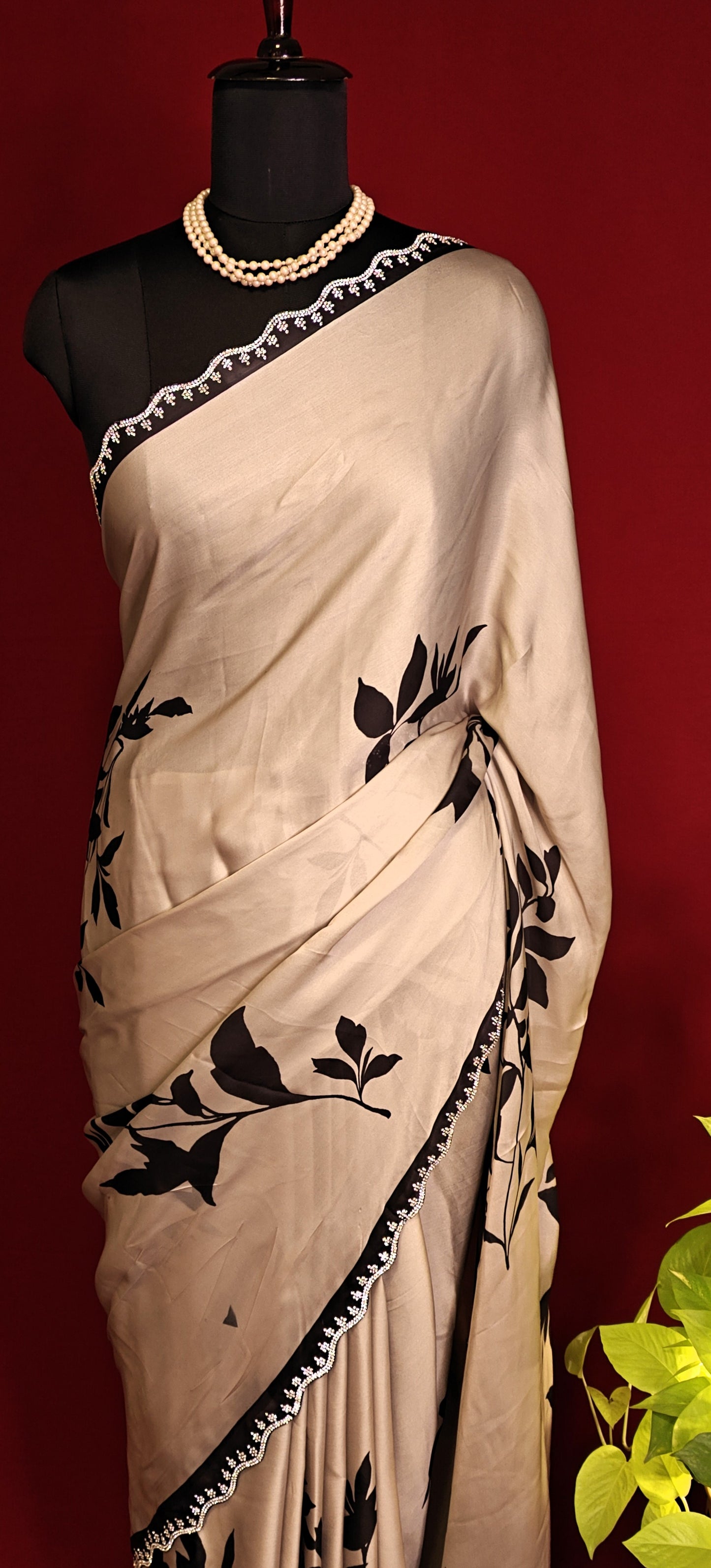 Cream satin georgette saree with medium-sized black leaf print, black motif border, and matching blouse piece on a mannequin against a red background