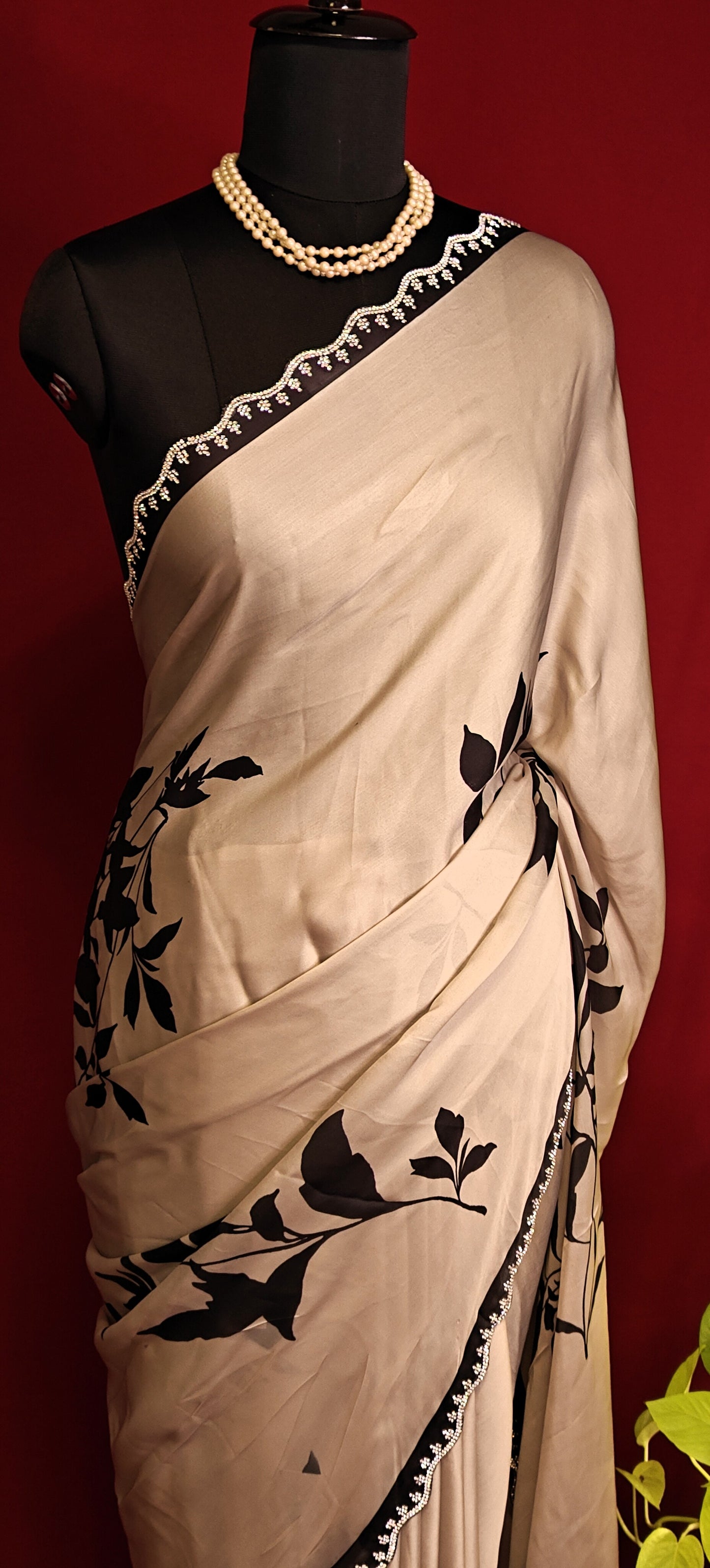 Cream satin georgette saree with medium-sized black leaf print, black motif border, and matching blouse piece on a mannequin against a red background