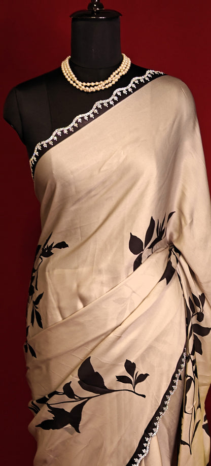 Cream satin georgette saree with medium-sized black leaf print, black motif border, and matching blouse piece on a mannequin against a red background