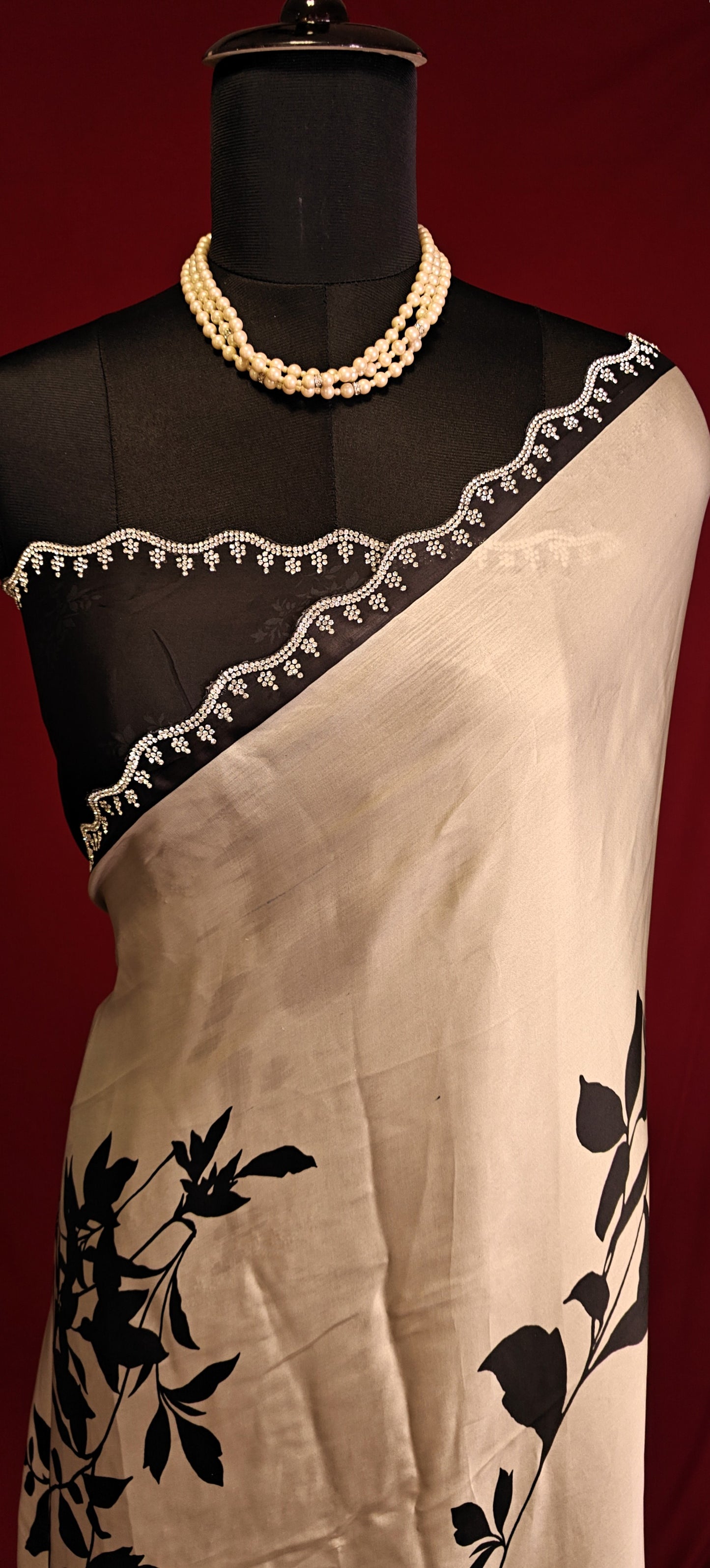 Cream satin georgette saree with medium-sized black leaf print, black motif border, and matching blouse piece on a mannequin against a red background