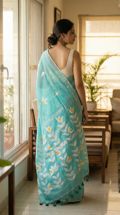 Woman in a sea green hand painted cotton linen saree with floral motifs and tassel pallu, standing in a well-lit room.