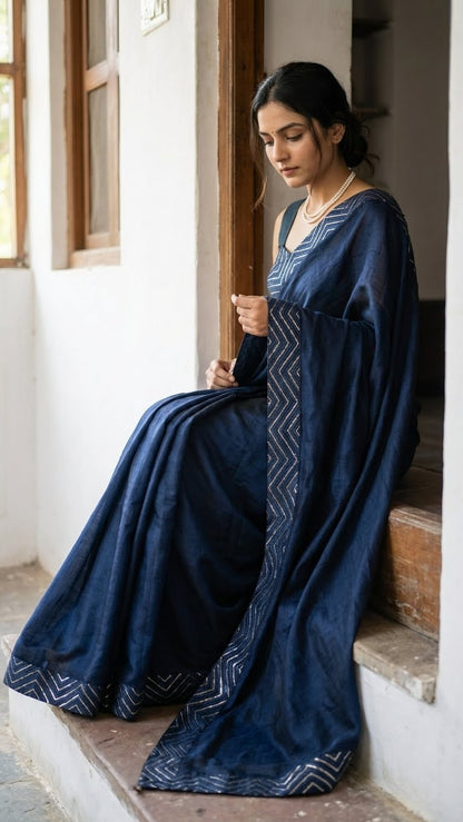 Woman in a blue silk saree with metallic zigzag border in gold and silver sitting on a step indoors.