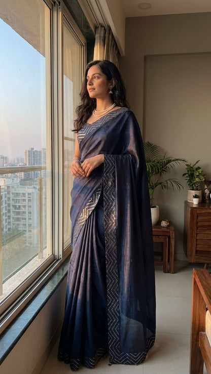 Woman in a blue silk saree with metallic zigzag border in gold and silver standing by a window with a cityscape view.
