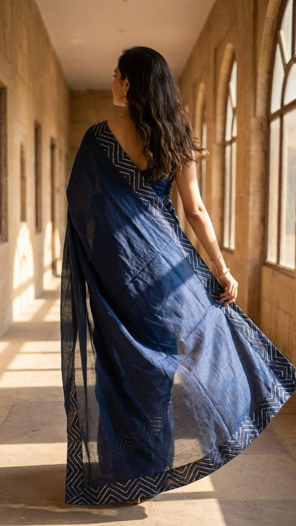 Woman in a blue silk saree with metallic zigzag border in gold and silver walking through a sunlit hallway.