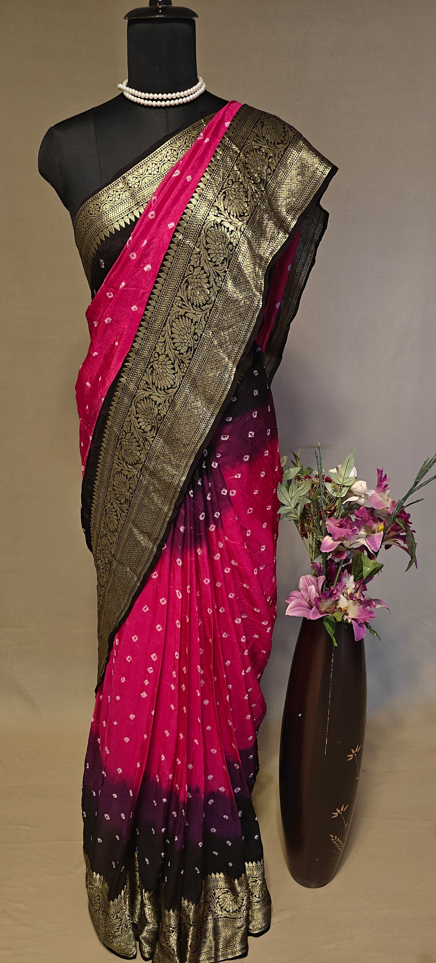 Magenta silk saree with broad golden zari border, styled on mannequin with a vase of flowers in the background.