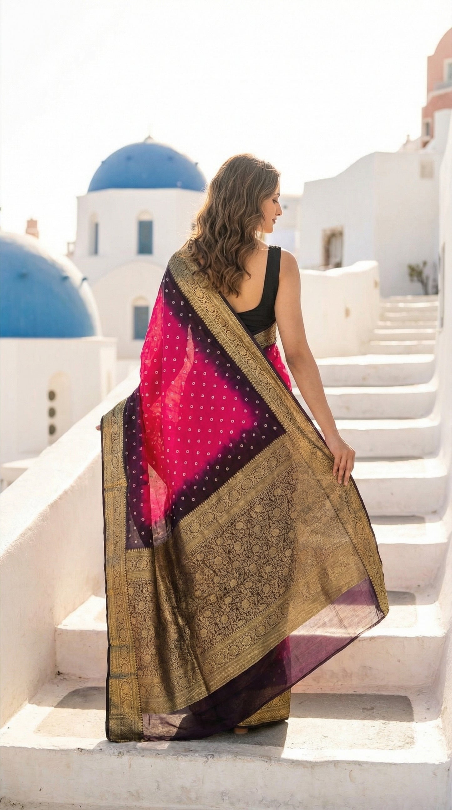 Woman in a Magenta silk saree with broad golden Zari border, standing on white steps with blue domes in the background.