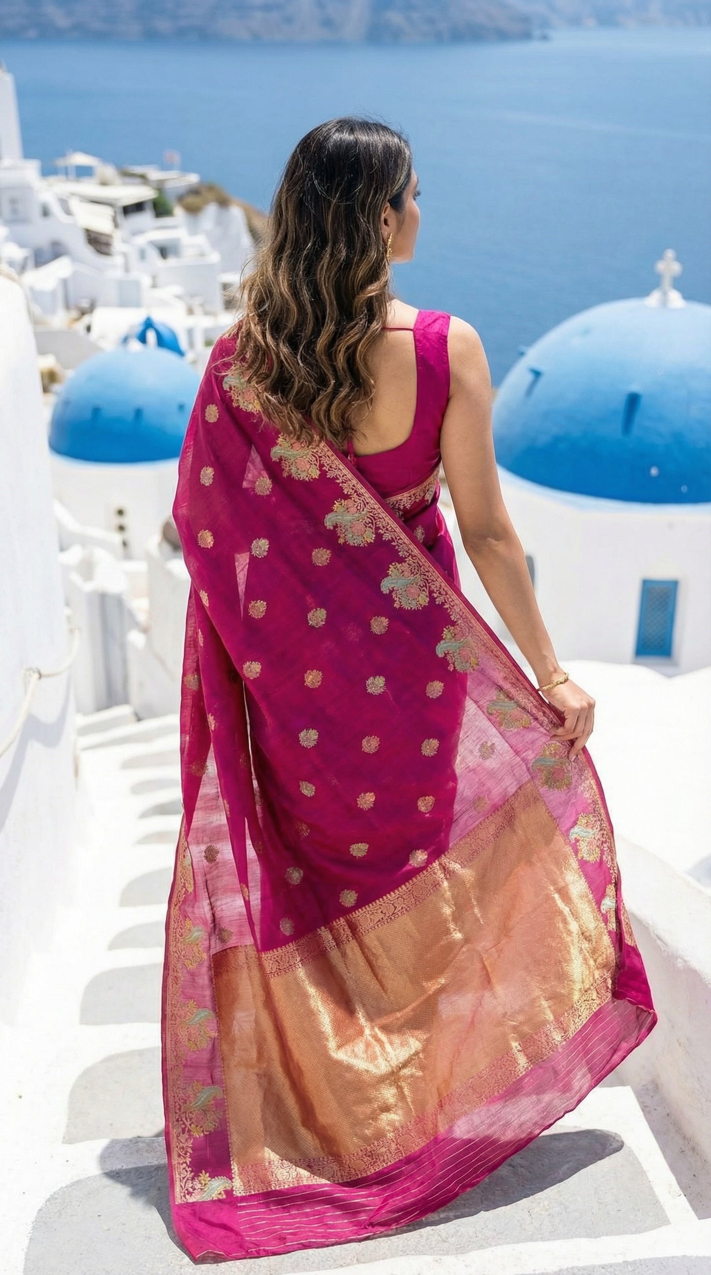 Woman in a Magenta Silk Saree with Zari Embroidery with parrot motifs, standing in a scenic location with blue domes and white buildings.
