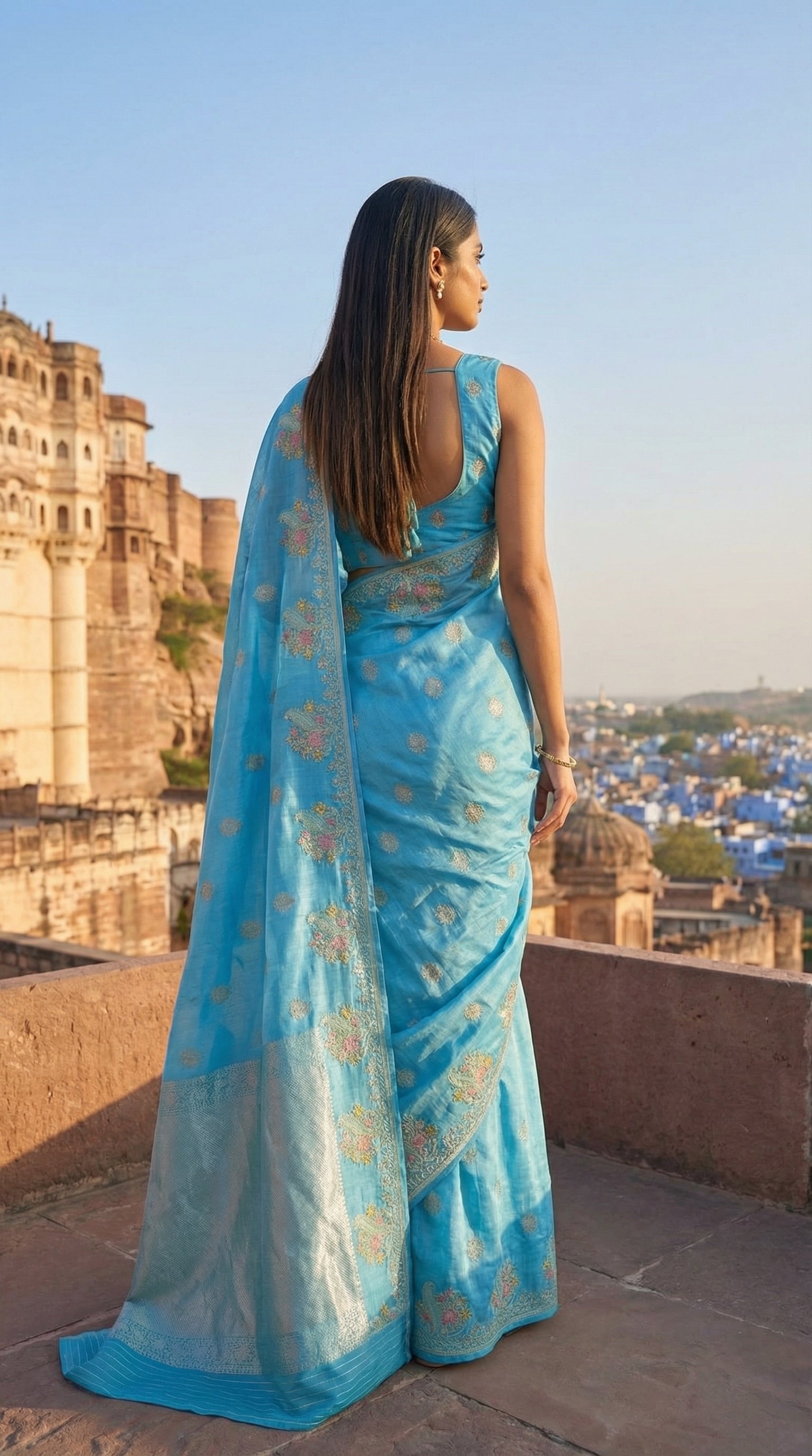 Woman in a Sky Blue Silk Saree with Zari Embroidery with parrot motifs, overlooking a cityscape with historical architecture.