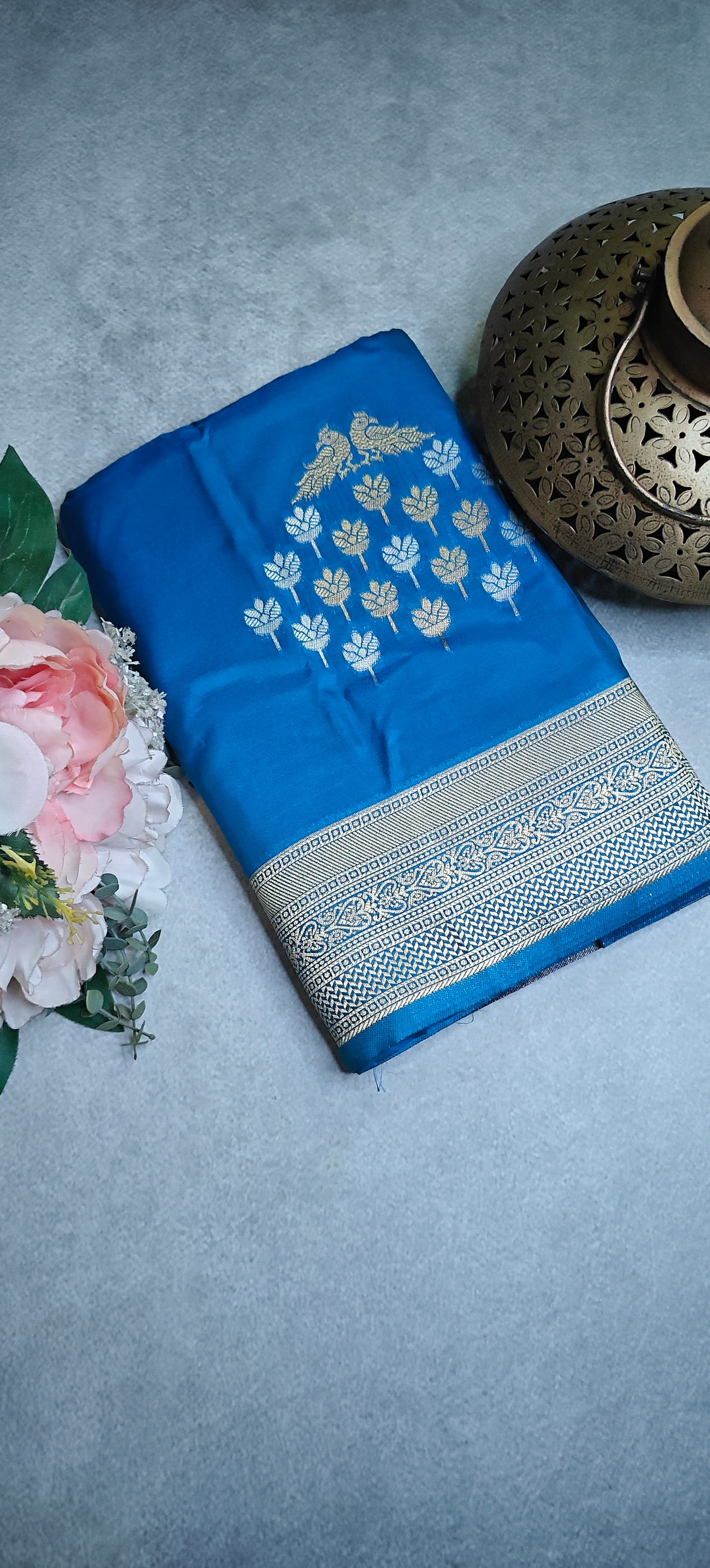 Blue soft silk saree with silver leaf motifs and luminous zari detailing on a gray surface with flowers.