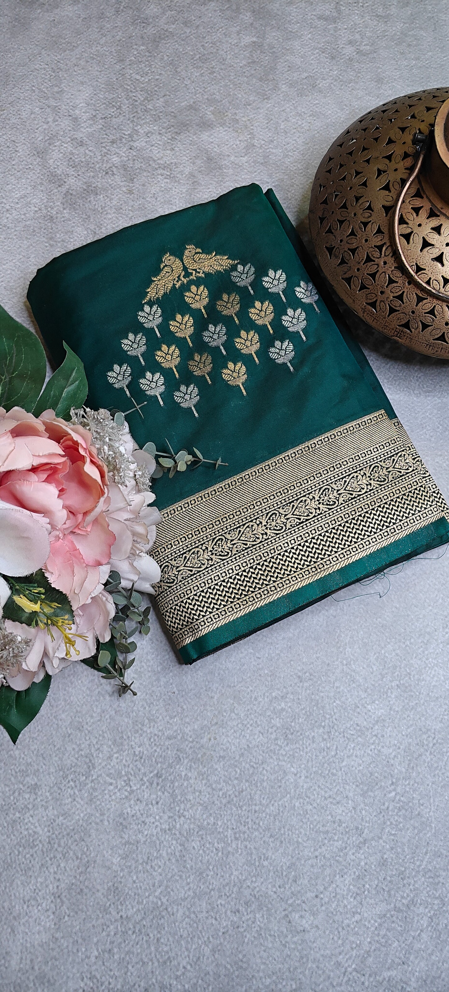 Green soft silk saree with bird and branch zari motifs and antique gold border on a gray surface with flowers and a decorative item.