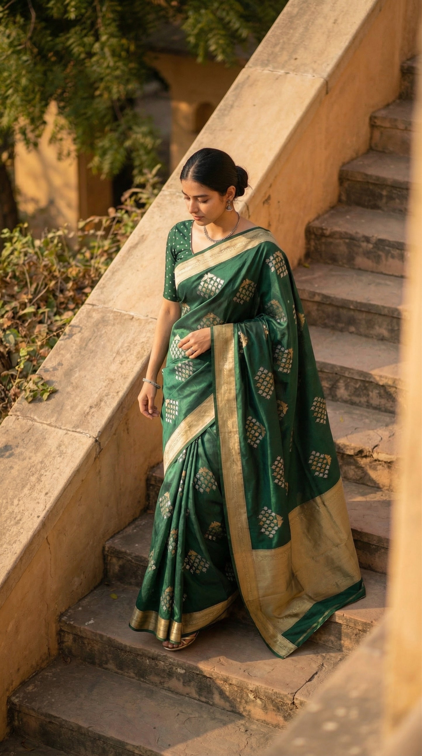 Woman wearing Green soft silk saree with bird and branch Zari motifs and antique gold border. Captured from a higher angle as she walks down stone stairs, showing the full length of the trailing pallu.