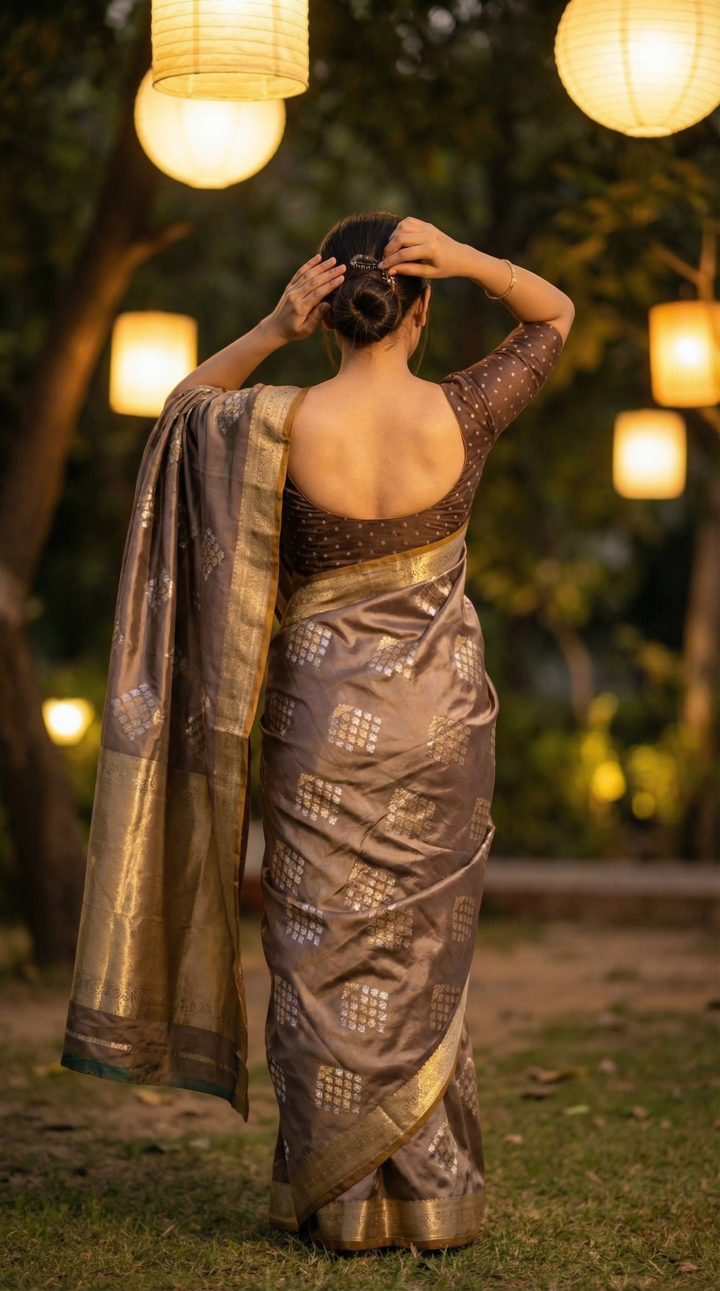 Woman wearing Woman wearing grey soft silk saree with golden embroidery and floral Zari border. ack-facing, both hands lifting to fix hair; highlights blouse back design + shoulder drape.