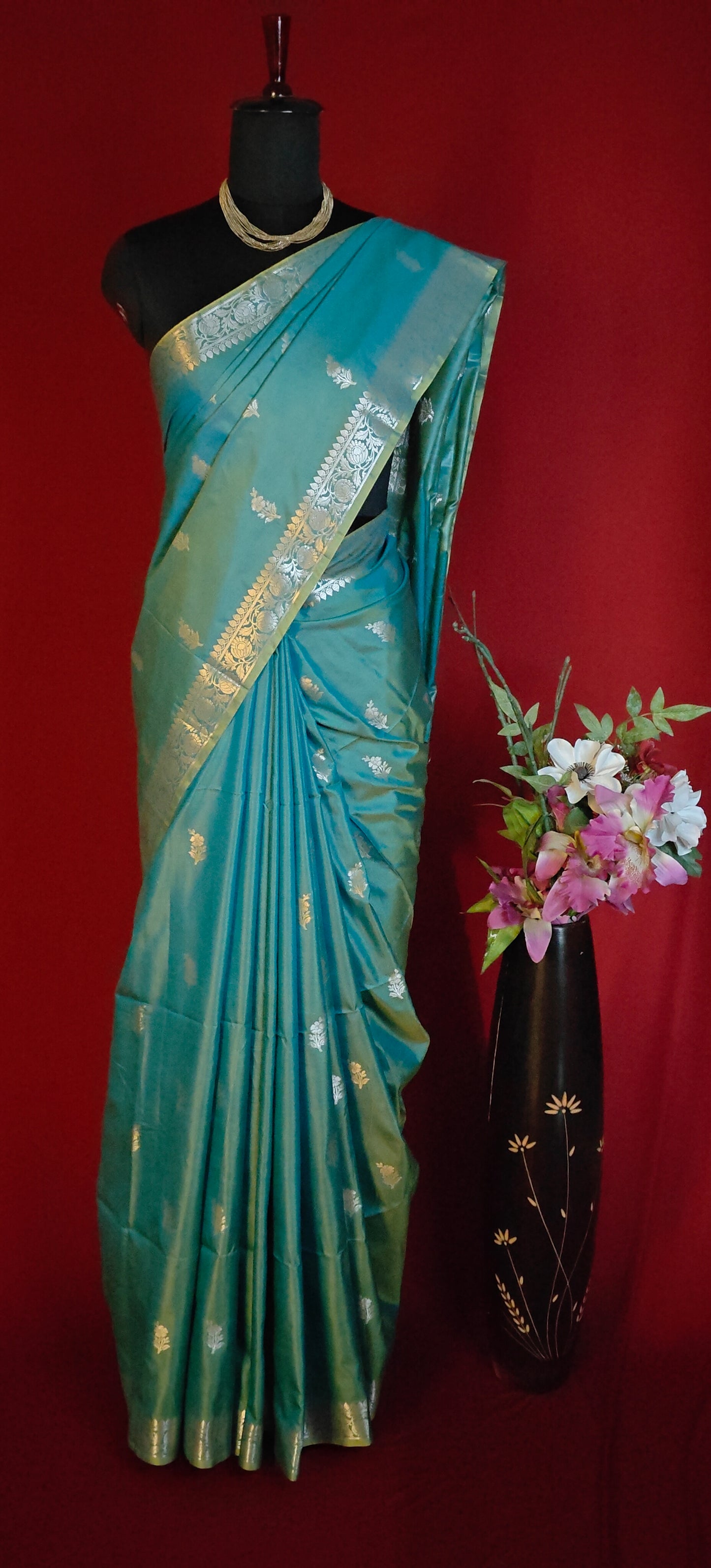 Cyan silk blend saree with woven floral motifs and traditional border with gold patterns on a mannequin against a red background.