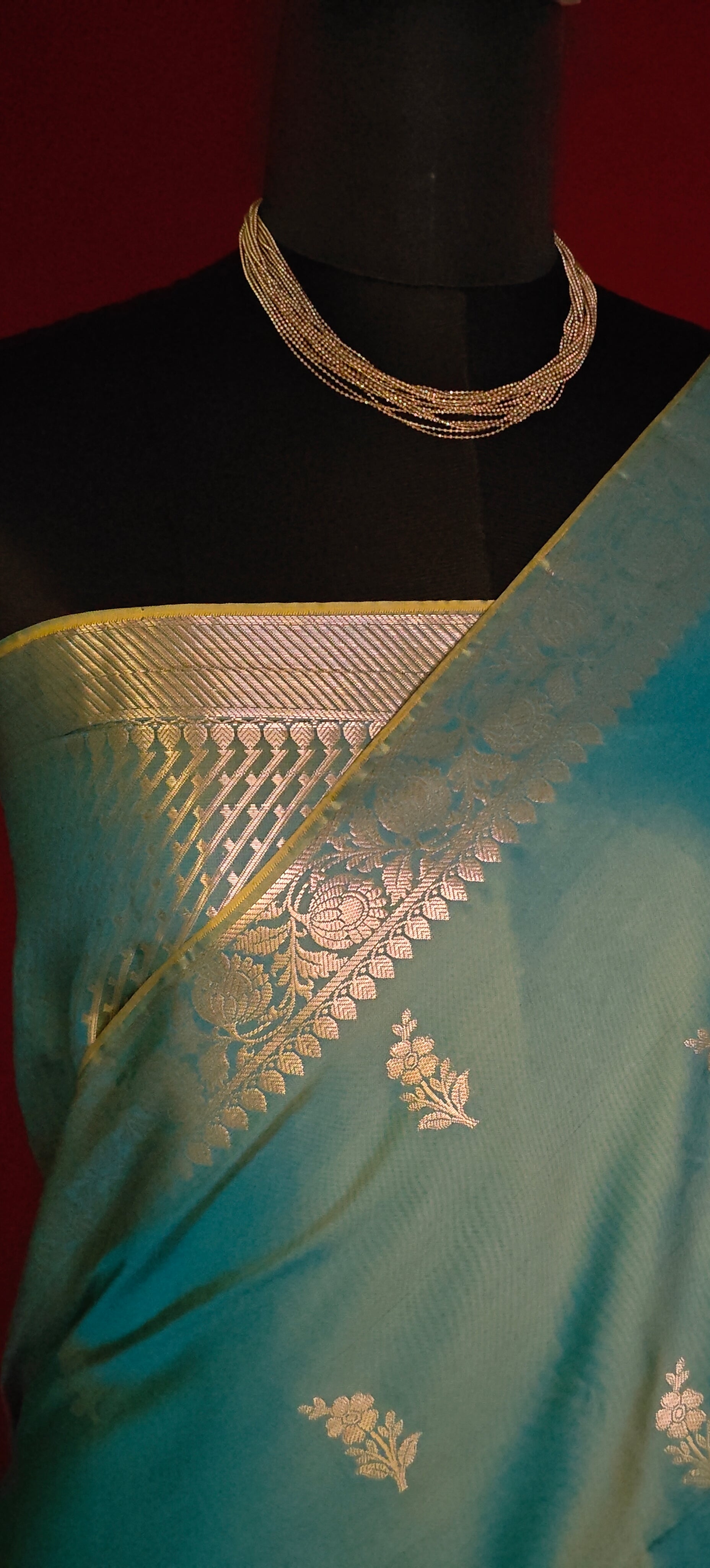 Cyan silk blend saree with woven floral motifs and traditional border with gold patterns on a mannequin against a red background.