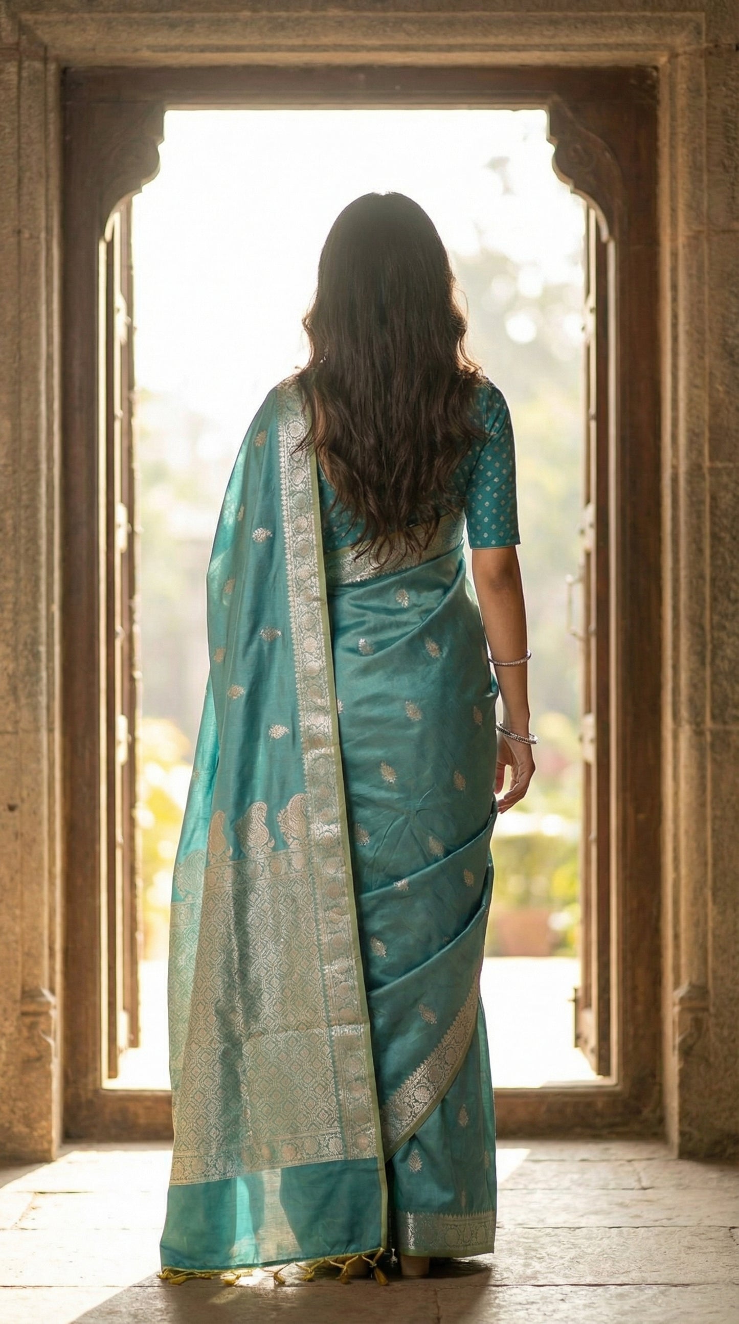 Woman wearing cyan silk blend saree with woven floral motifs and traditional border with gold pattern, back-facing — Castle doorway silhouette: Bright outside light, halo edges.