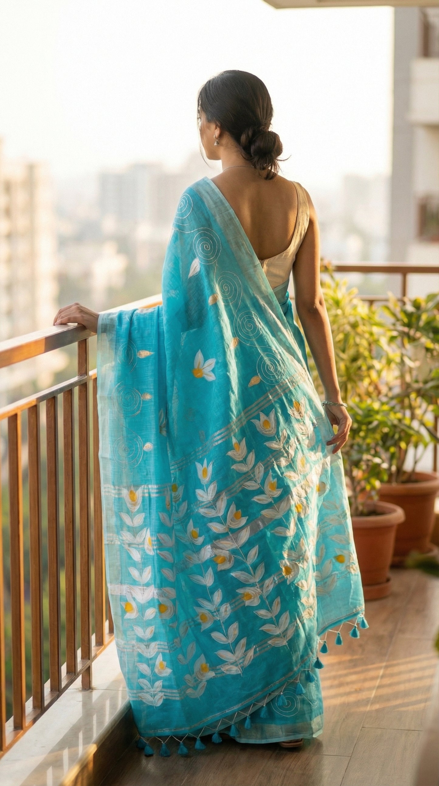 Woman in a sky blue hand painted cotton linen saree with floral motifs and tassel pallu, standing on a balcony.