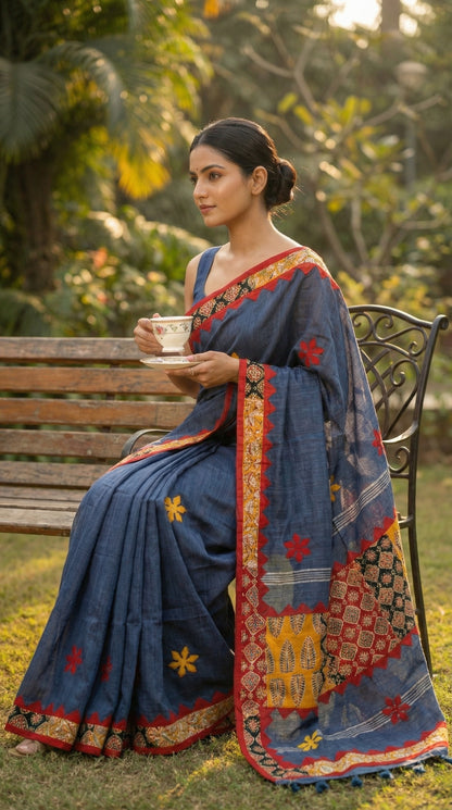 Woman in a slate grey linen cotton saree with traditional applique border, sitting outdoors holding a cup.