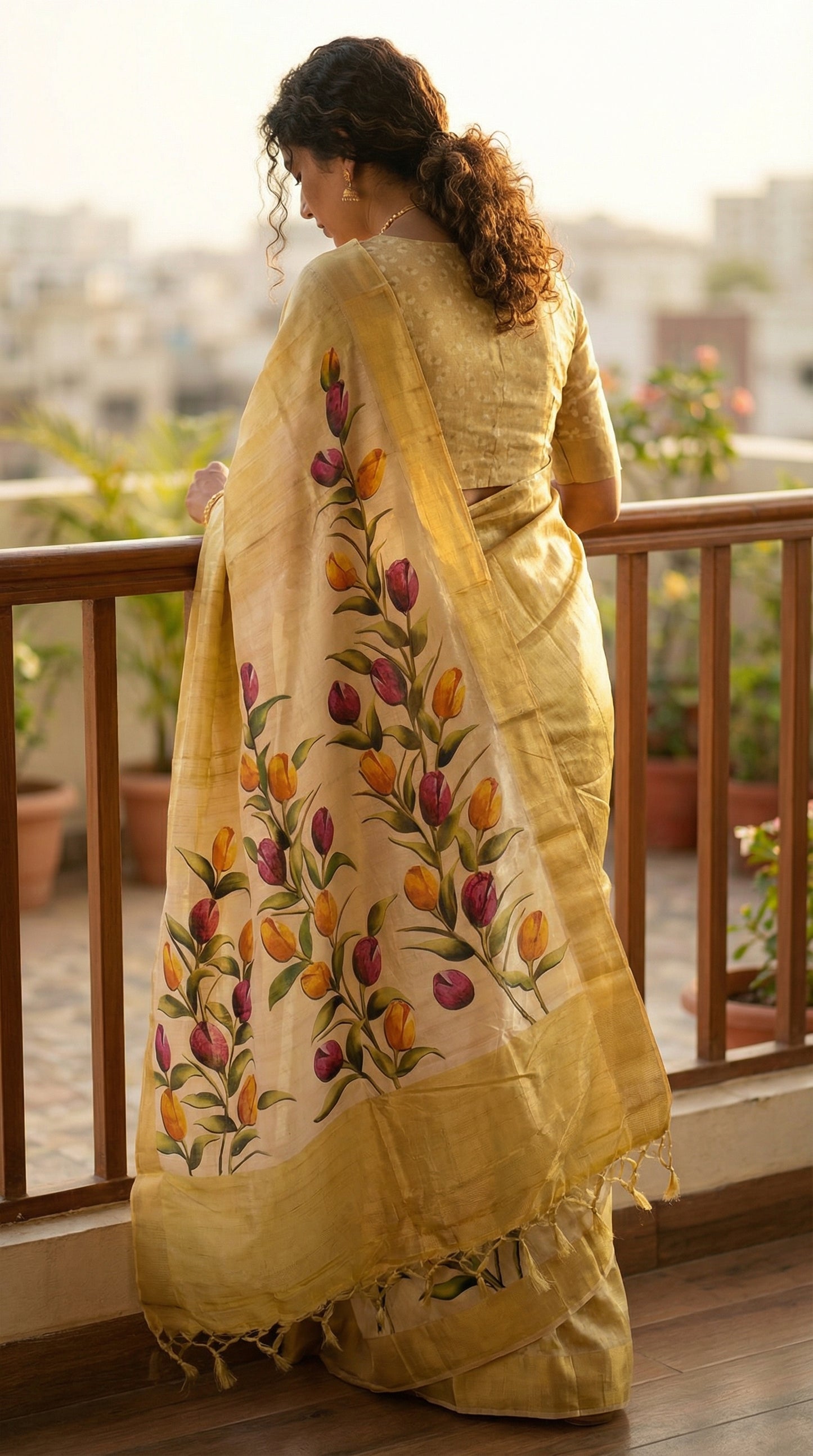 Woman wearing Golden viscose silk saree with subtle sheen and botanical motifs, festive Indian ethnic wear. Leaning forward on a railing with the back to the camera; the pallu hangs vertically, showing the full length of the border design.