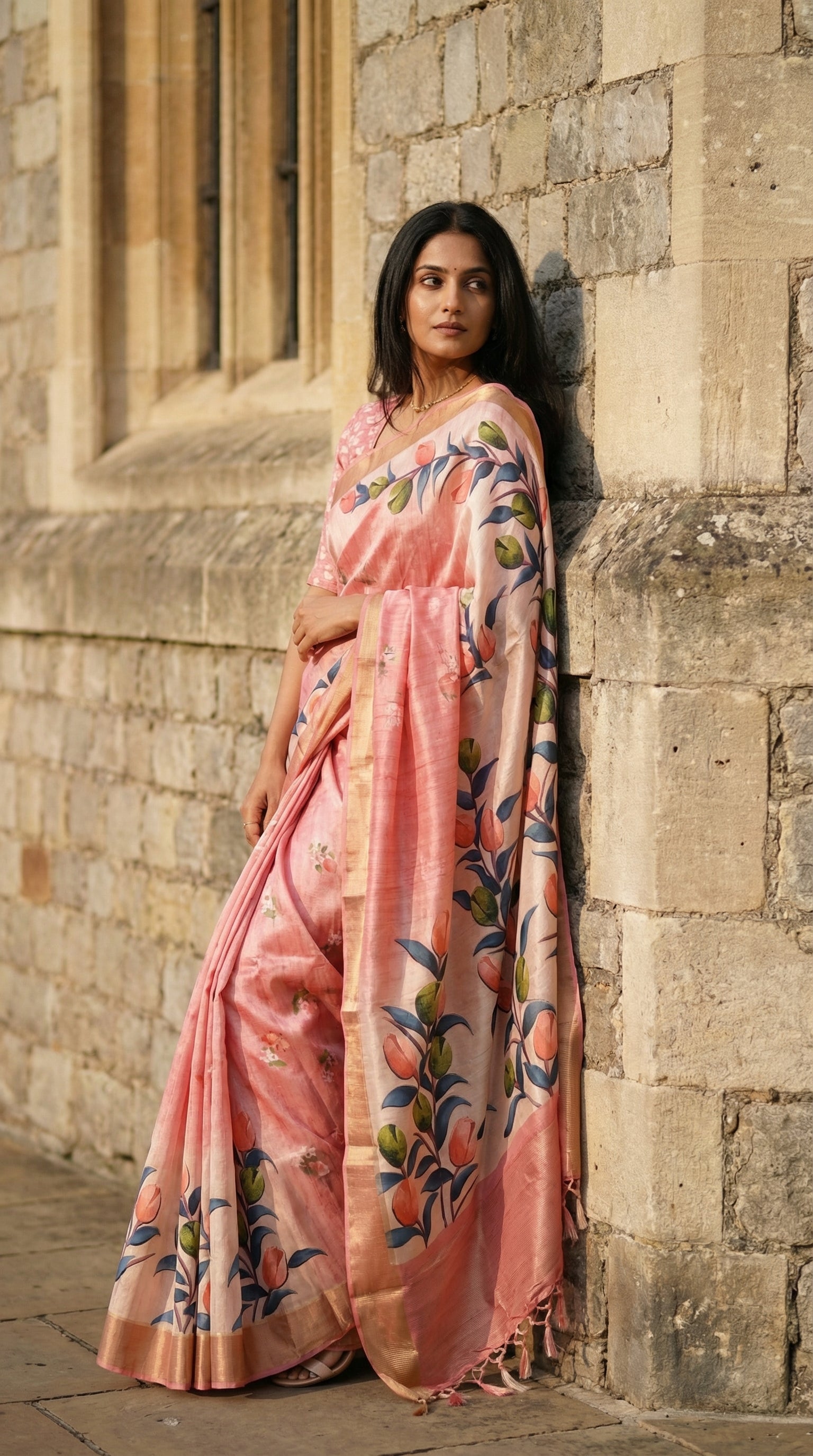Woman wearing Pink viscose silk saree with floral accents, leaning her side against an old palace wall (England's royal Windsor Castle), looking away from the camera with a faraway, mysterious expression.