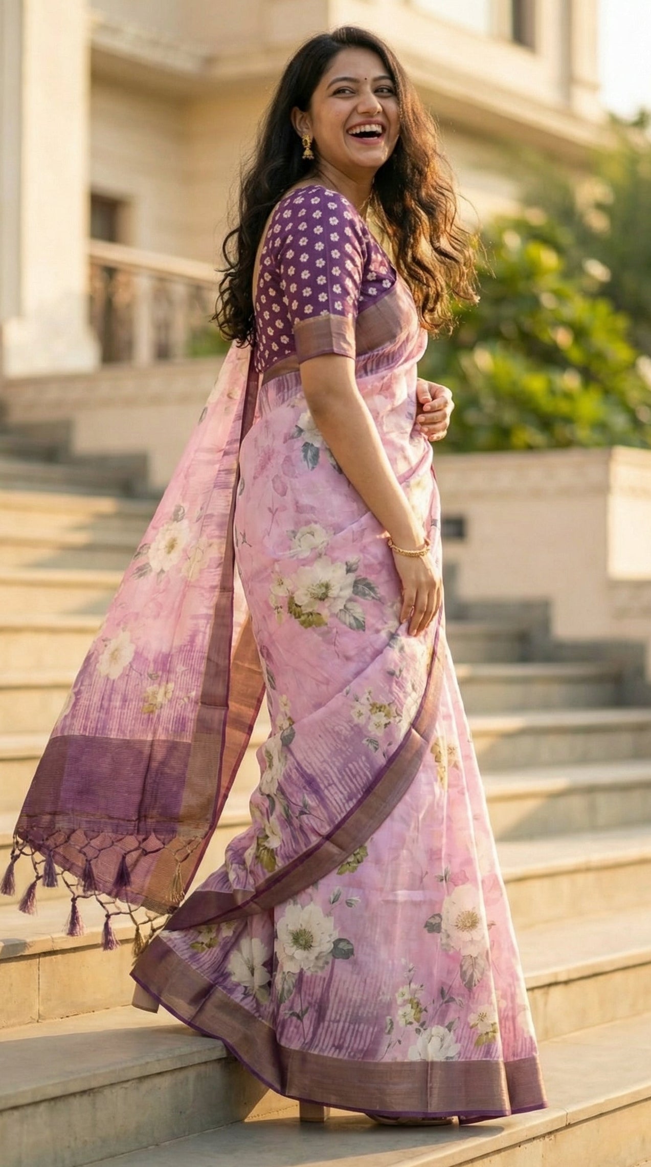 Woman wearing Lavender viscose silk blend saree with soft floral motifs, she is walking up a set of grand stairs, looking back while laughing, showing a beautiful spiral drape of the saree from the side.