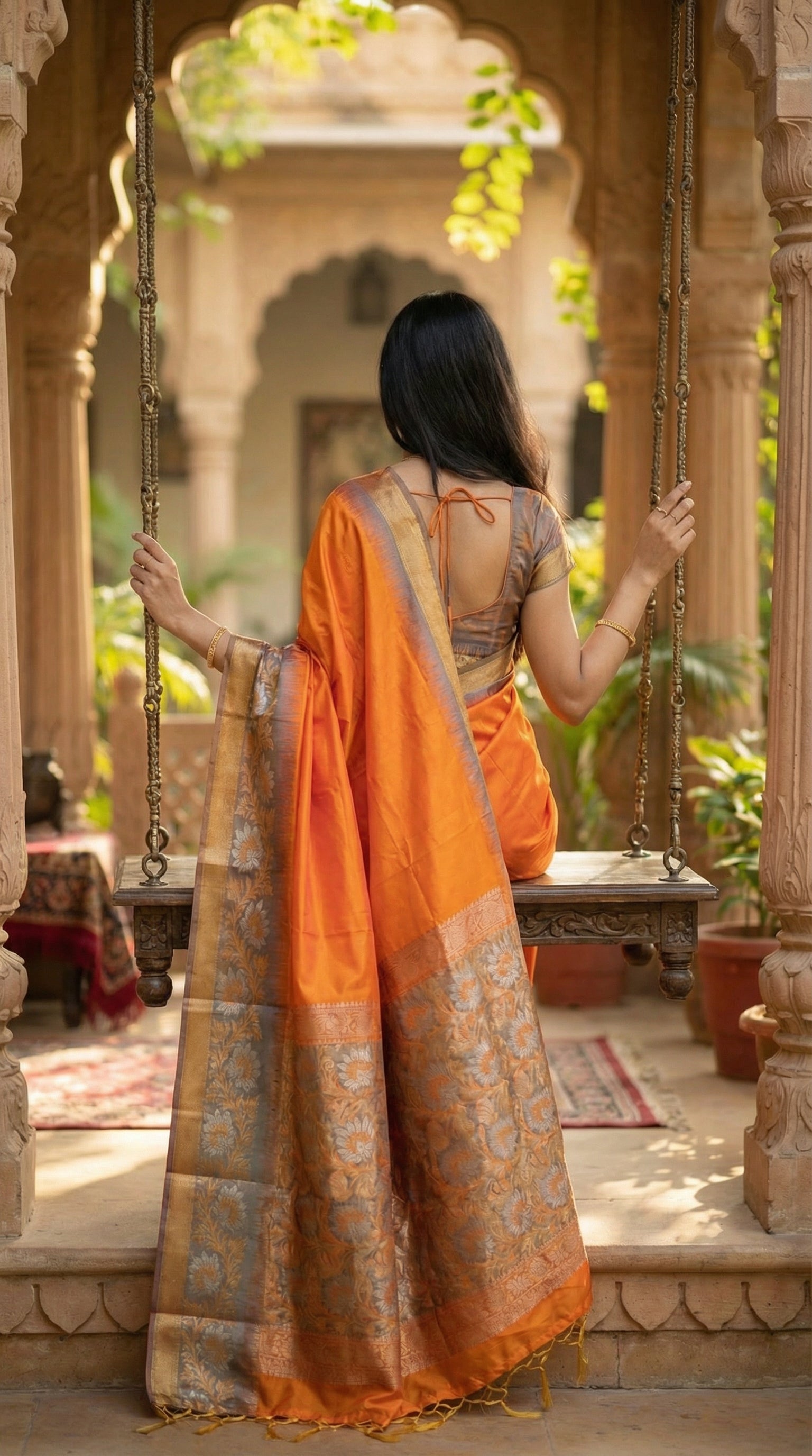 Woman in an Orange viscose silk saree with copper brown border and floral embroidery, elegant festive ethnic wear, sitting on a swing in a traditional setting.