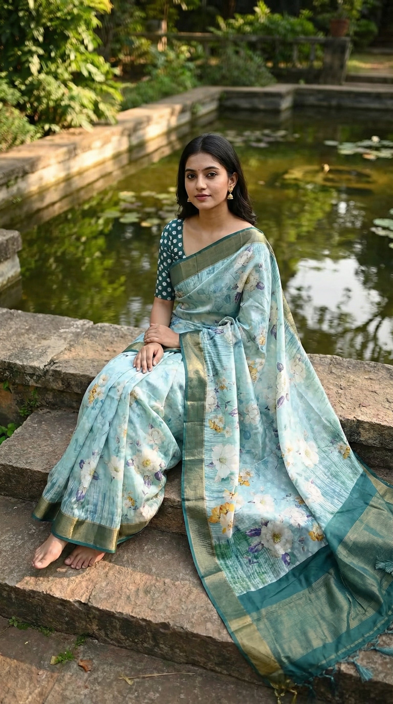 Woman wearing Sea green viscose silk saree with floral print, sitting comfortably near a pond in a garden.
