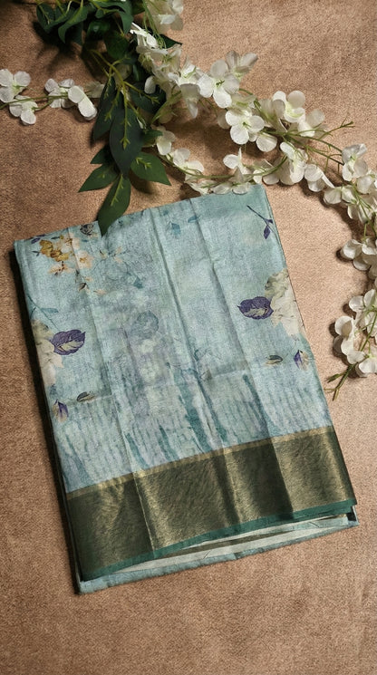 Sea green viscose silk saree with floral print lying on a flat surface with flowers.