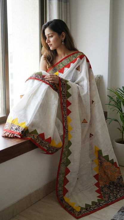 A lady wearing White cotton-linen saree with colorful red, green, and yellow triangle appliqué and printed borders sitting on a window in her house.