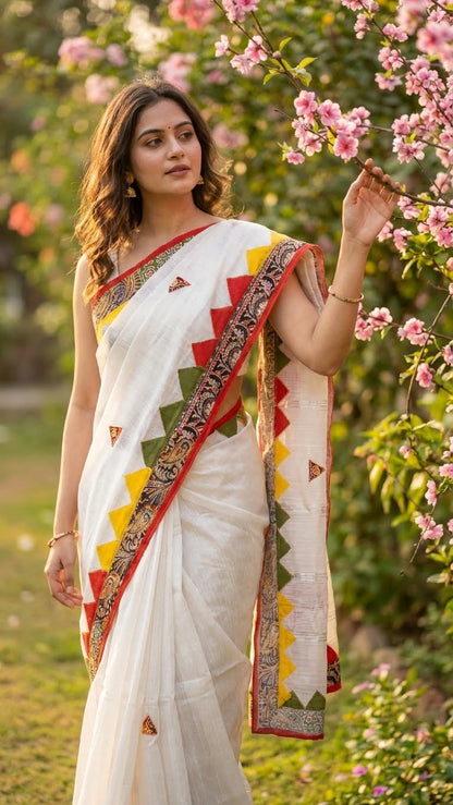 A lady wearing White cotton-linen saree with colorful red, green, and yellow triangle appliqué and printed borders standing and touching flowers in a garden.