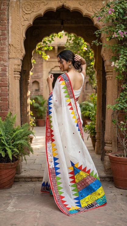 Woman in a white cotton saree with vibrant multi-color triangle appliqué and geometric printed borders standing in front of an archway with plants.