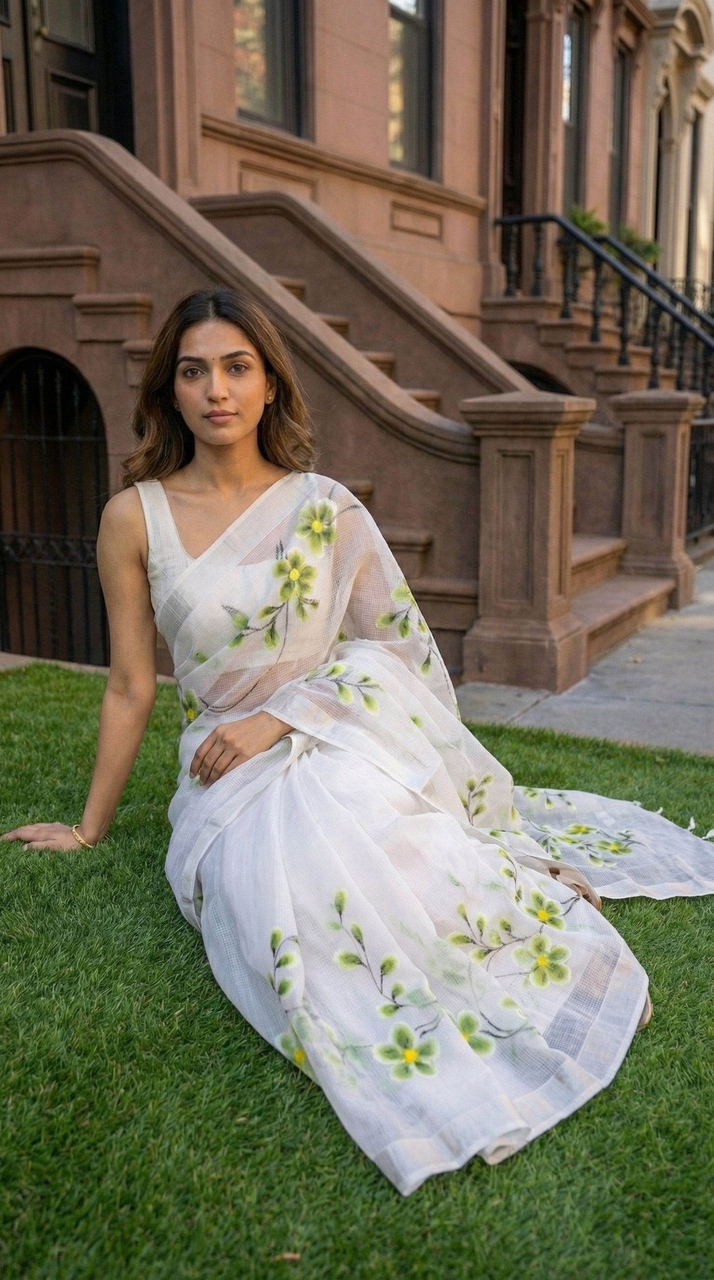 Woman in a white-green-floral-kota-doria-saree, sitting on grass in front of a building.
