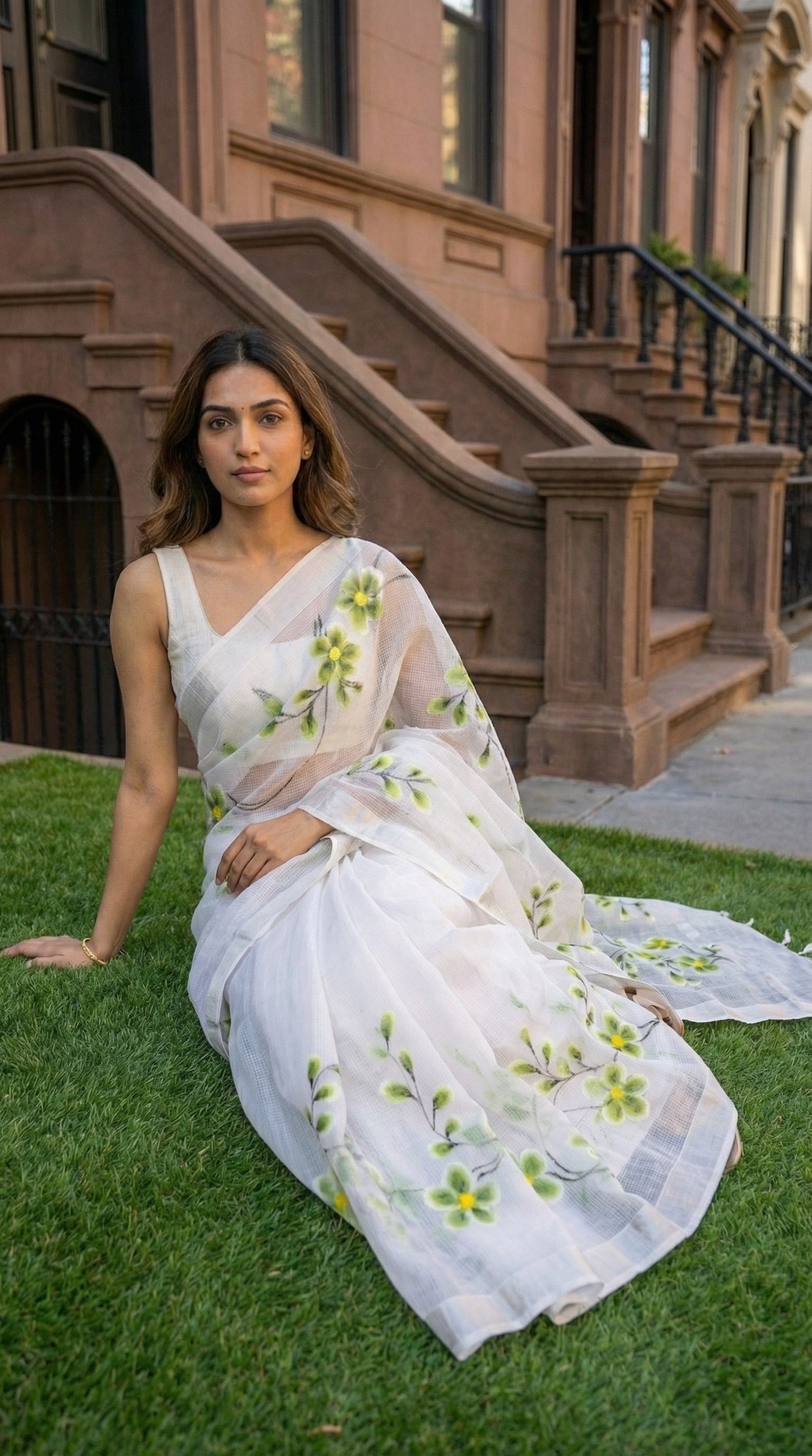 Woman in a white-green-floral-kota-doria-saree, sitting on grass in front of a building.