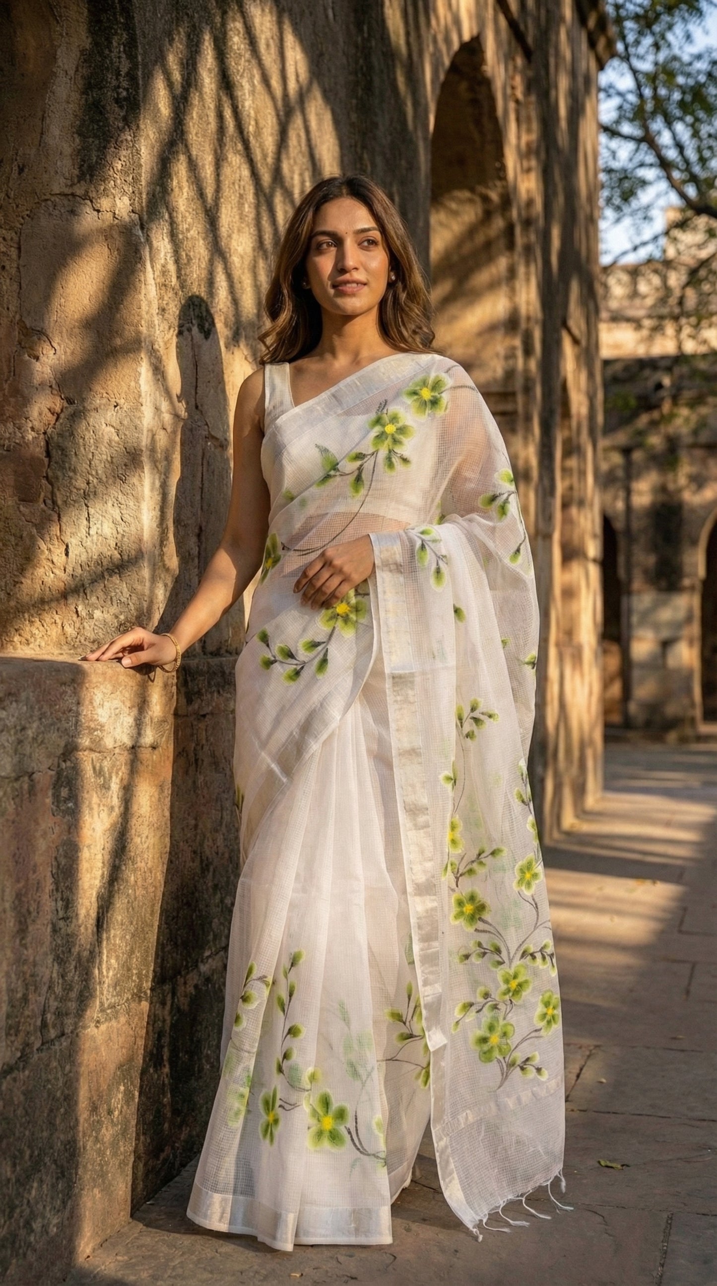 Woman in a white-green-floral-kota-doria-saree, standing against a stone wall.