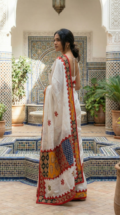 Woman in a white linen cotton saree with traditional applique border and heritage detailing, standing in a decorative indoor setting.