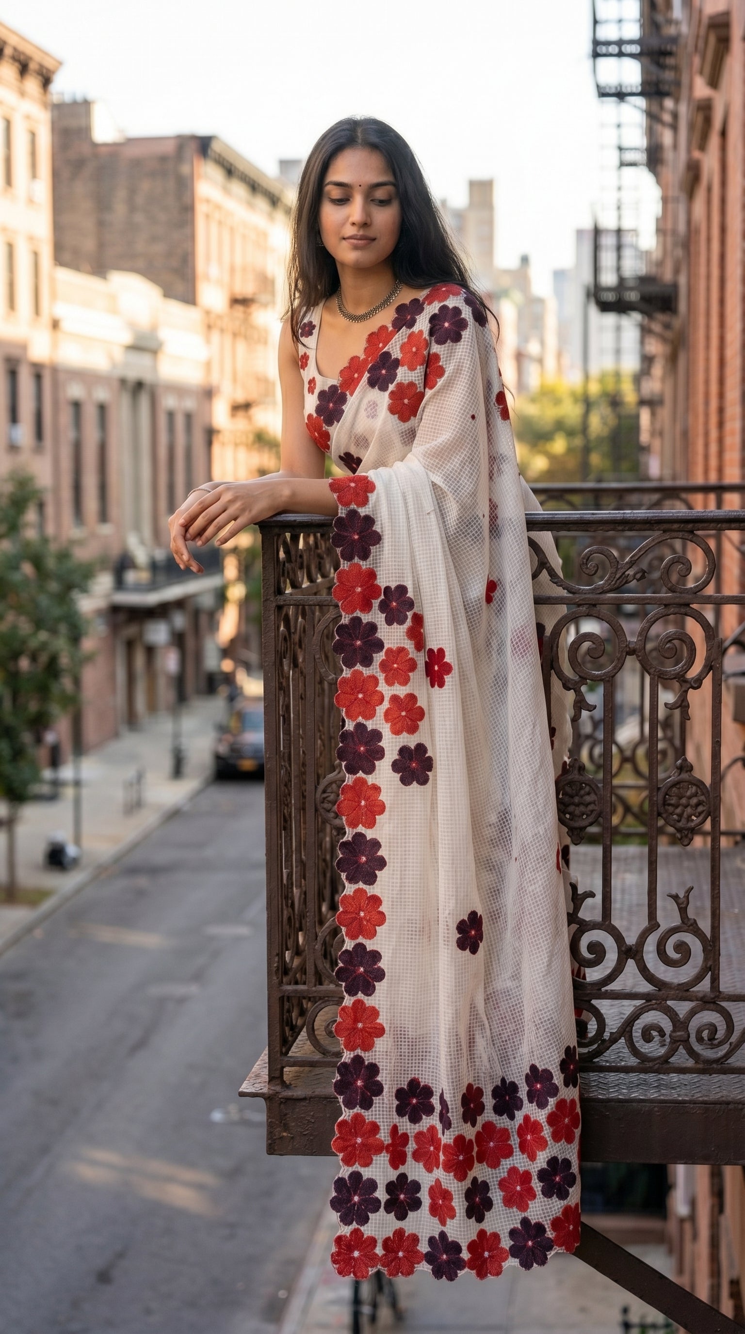 Woman in a white-maroon-brown-mirror-saree, standing on a balcony with a city street in the background