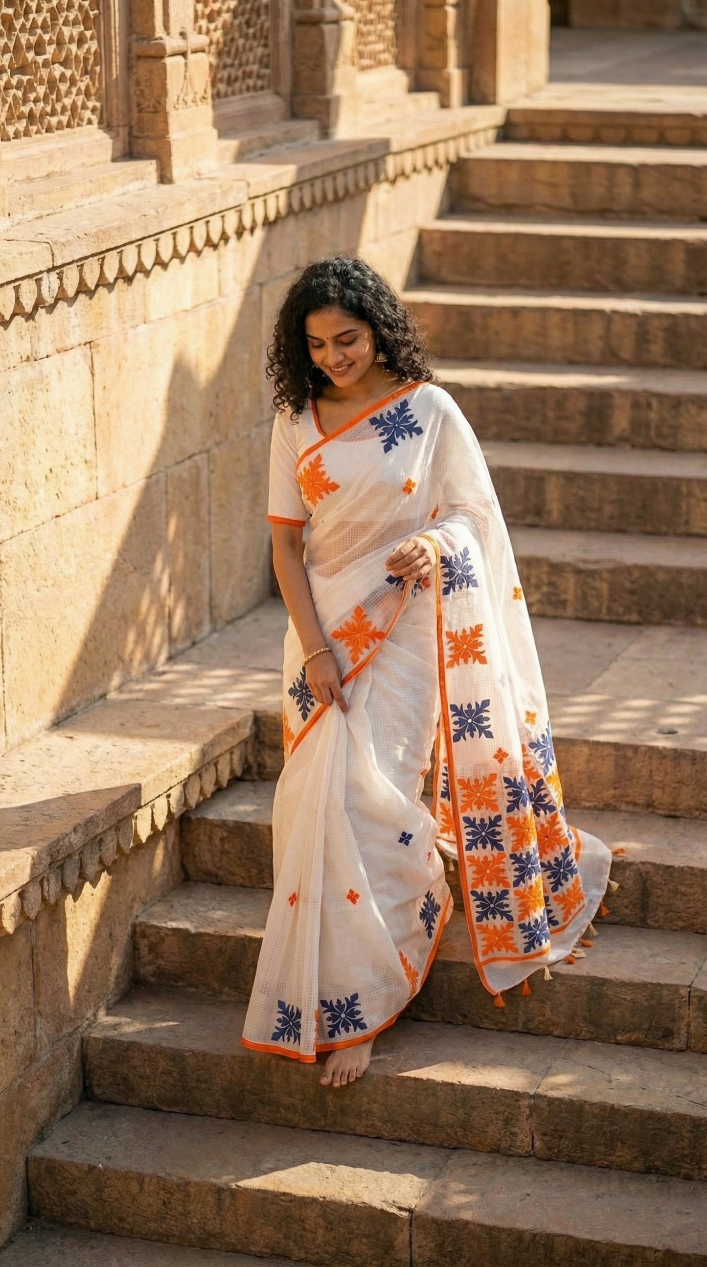 Woman in a white-orange-grey-applique-kota-saree, standing on stone steps with architectural background