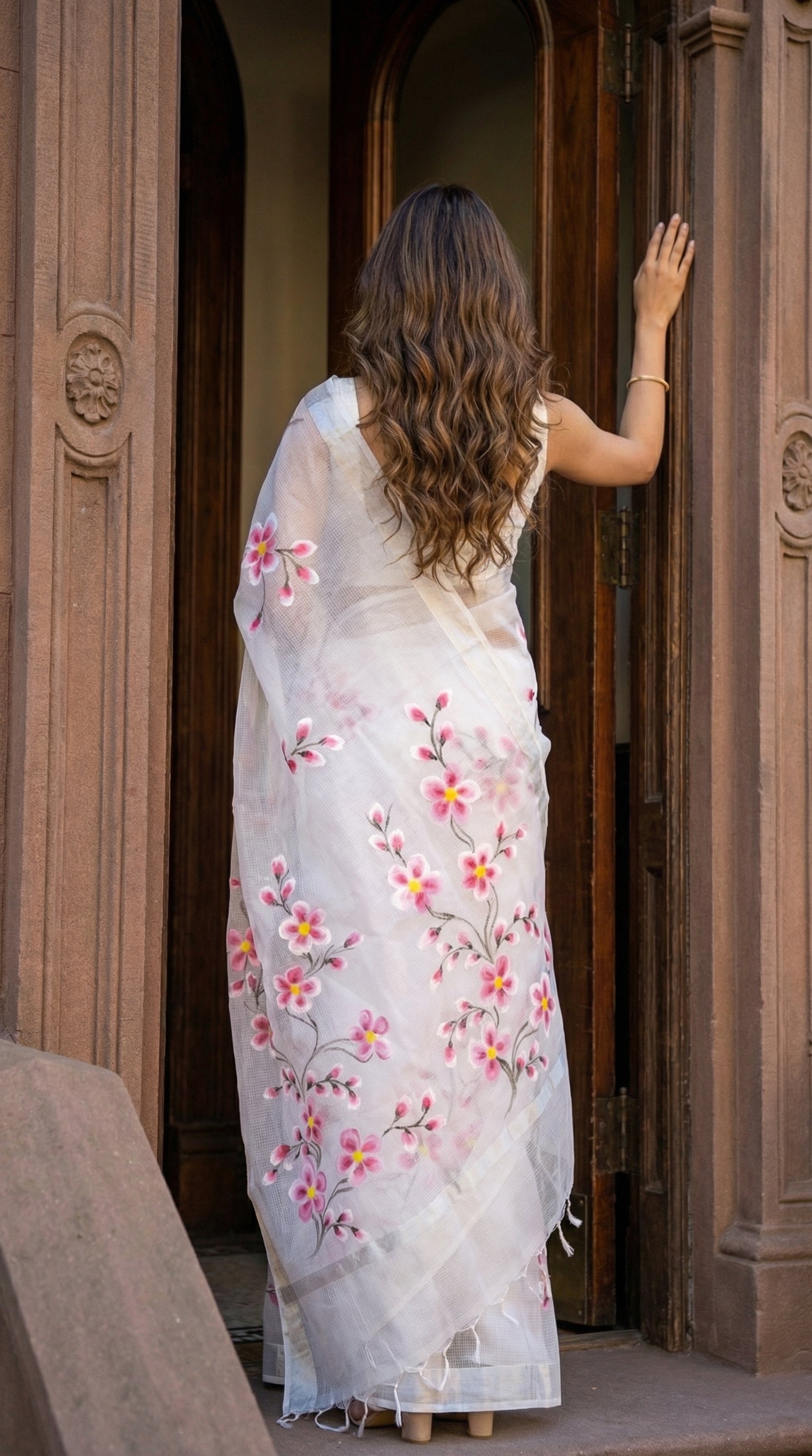 Woman wearing a white-pink-floral-kota-doria-saree, standing in front of a wooden door.