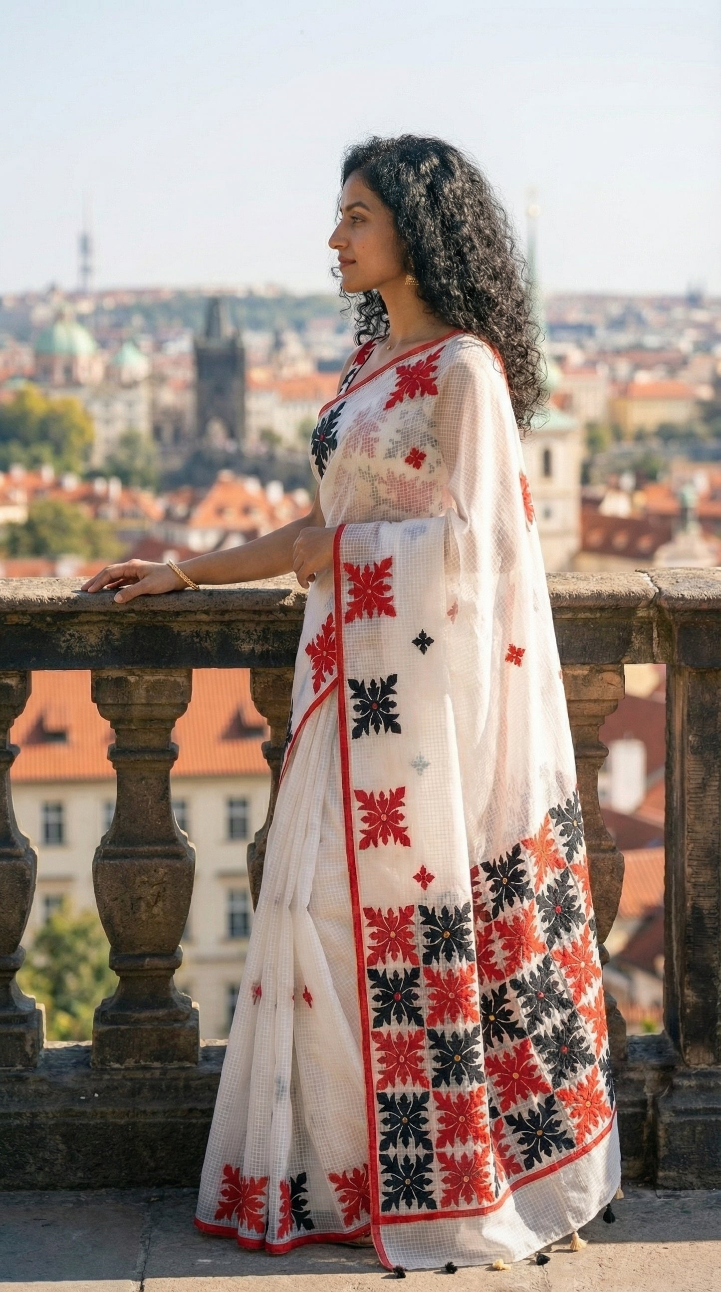 Woman in a white-red-black-applique-kota-saree, against a colorful tiled wall. standing on a balcony with a cityscape in the background