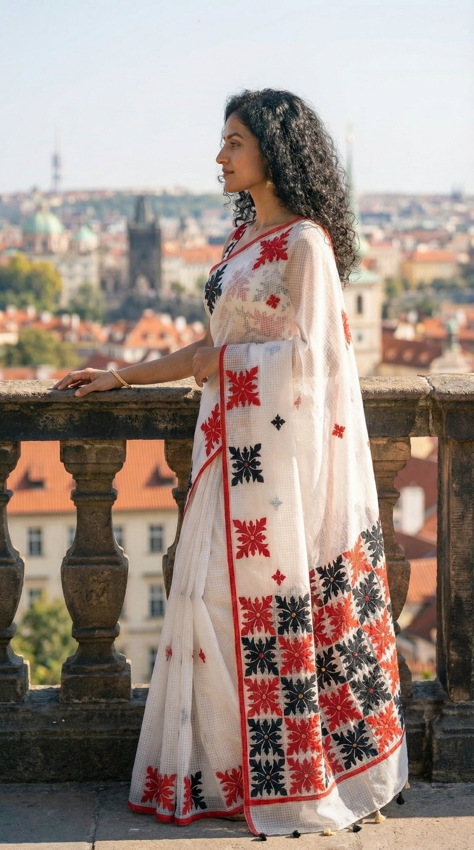 Woman in a white-red-black-applique-kota-saree, against a colorful tiled wall. standing on a balcony with a cityscape in the background