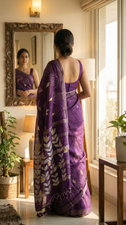 Woman in a wine hand painted cotton linen saree with floral motifs and tassel pallu, standing in a room with a mirror and window.