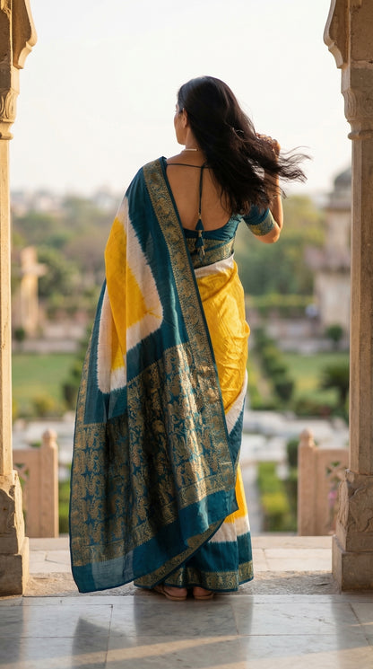 Woman wearing a yellow modal silk saree with teal Zari border and brocade pallu, standing in an outdoor setting with greenery.