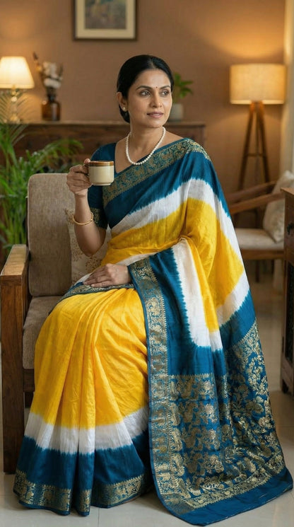 Woman wearing a yellow modal silk saree with teal Zari border and brocade pallu, holding a cup in a cozy indoor setting
